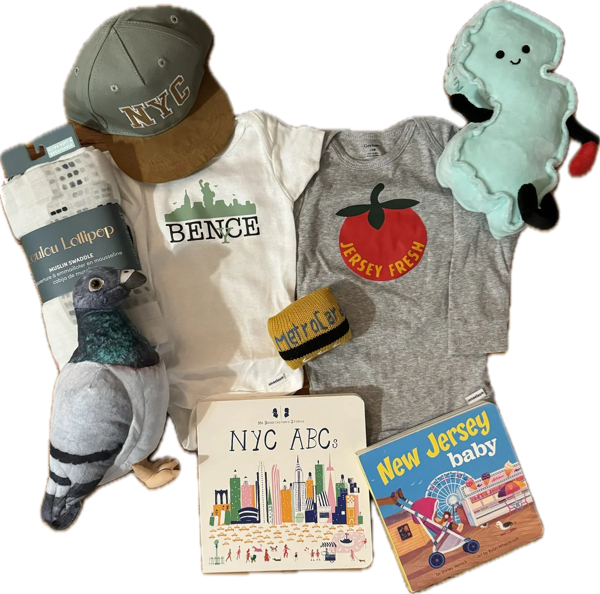 A new-baby gift assembled as one bundle: a custom bodysuit printed with a name and the New York skyline, a Jersey Fresh tomato bodysuit, an NYC cap, a plush pigeon, a plush New Jersey, a MetroCard-patterned hat, a muslin swaddle, and two picture books.